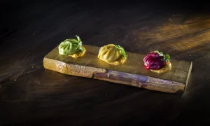 hree colorful dumplings arranged in a row on a rustic wooden board, each sitting on a small dollop of sauce—one green, one golden-yellow, and one deep red—garnished with herbs, against a dark wooden background